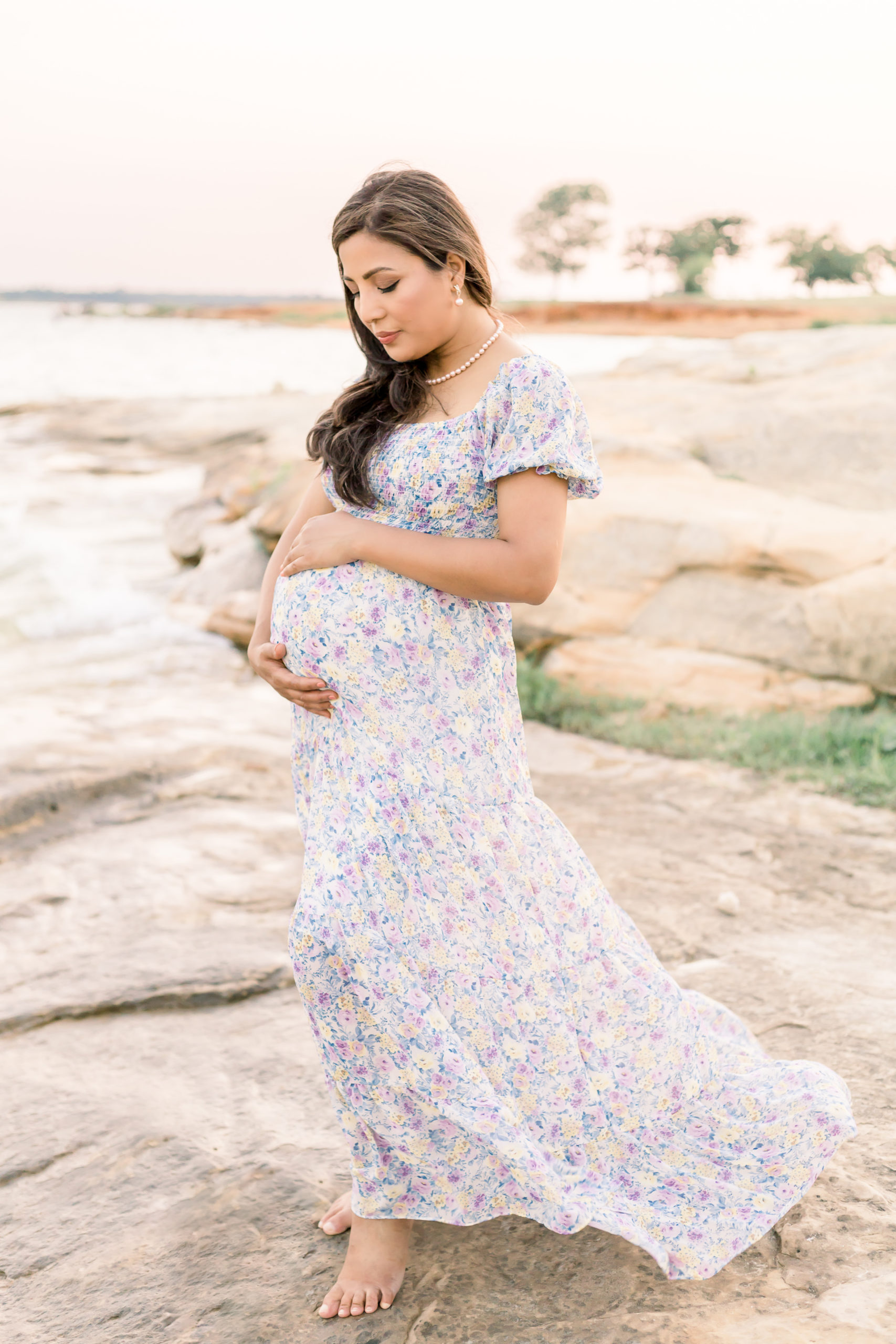Pregnant woman at Murrell Park posing on the rocks by lake grapevine for her maternity portraits in Flower Mound wearing a long flowy maxi dress