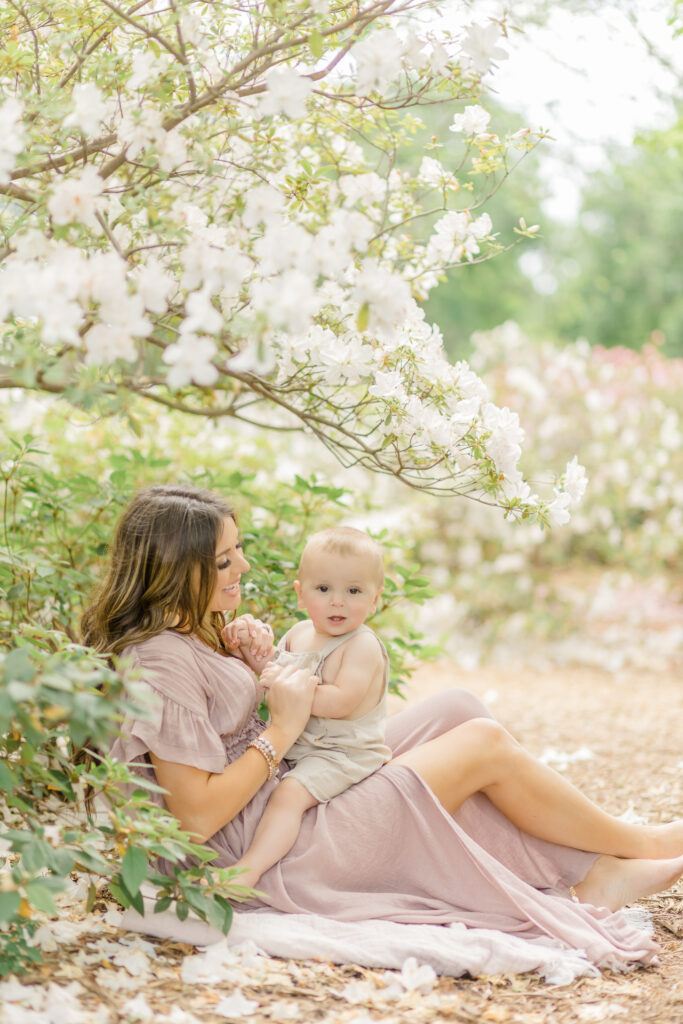 Mother and baby son holding hands playfully during a spring portrait session at the dallas arboretum surrounded by white flowers wearing a light pink dress 